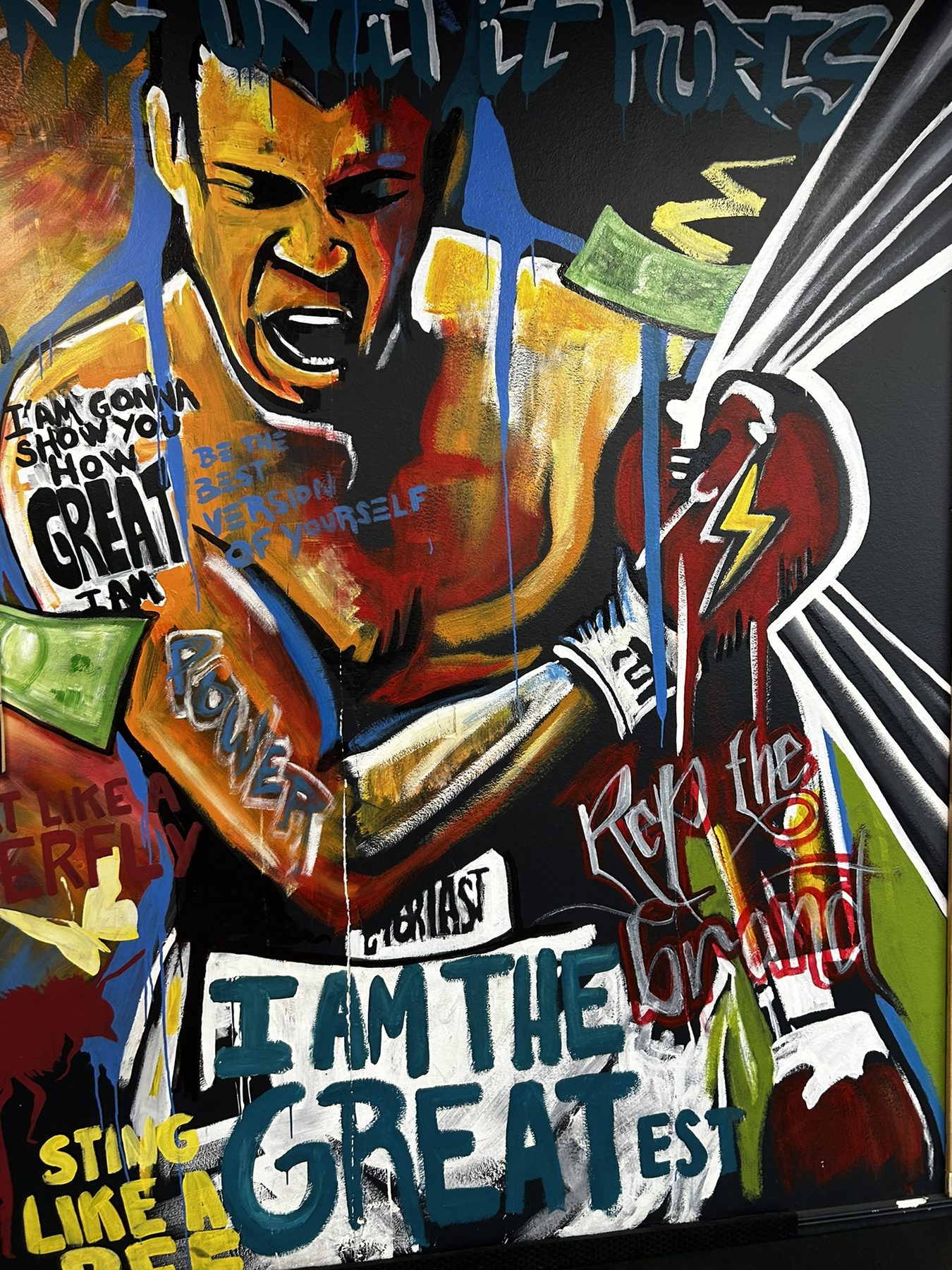 Hand-painted expressionist mural of a young Muhammad Ali mid-shout, surrounded by hand-lettered phrases including 'I AM THE GREATEST,' 'Sting Like a Bee,' and 'Float Like a Butterfly'